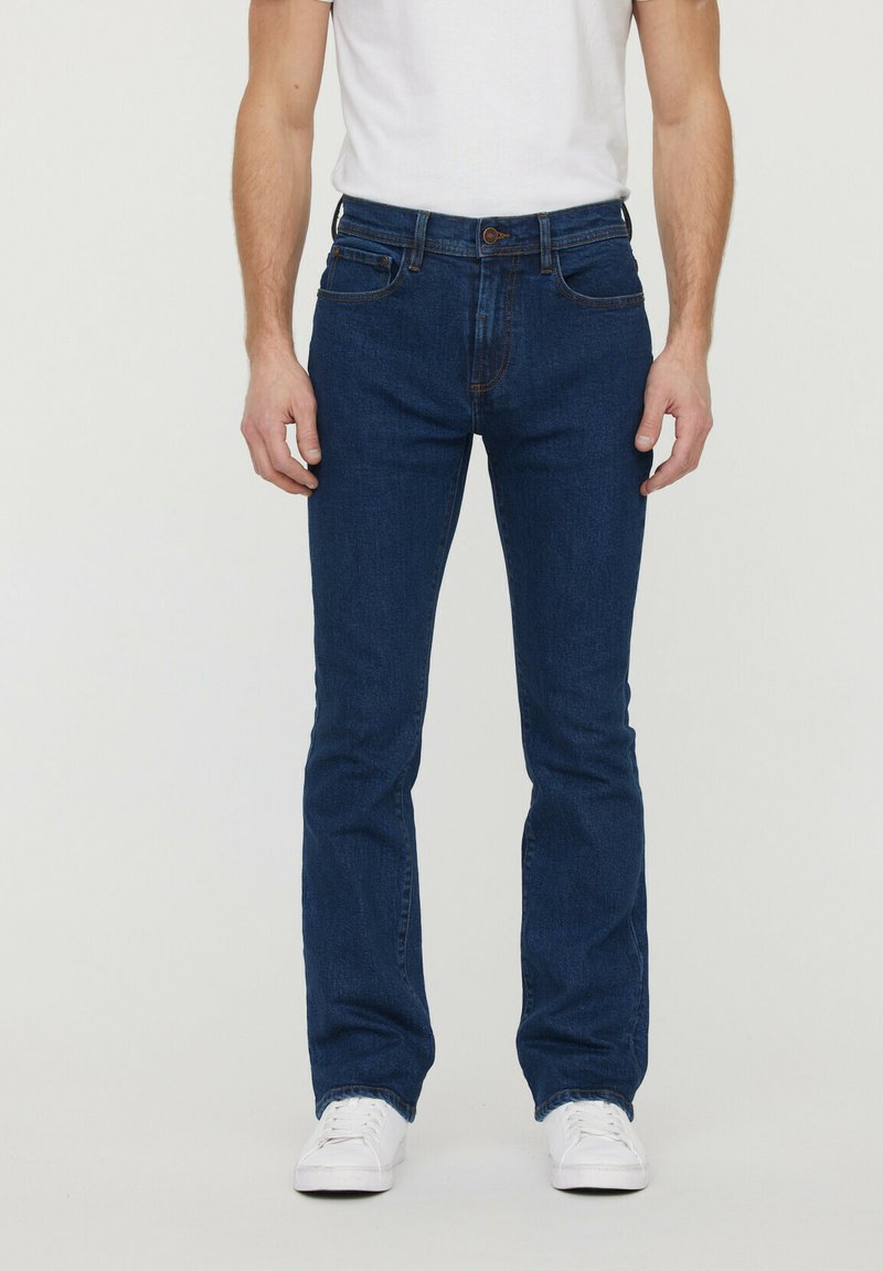 Jean en denim bleu foncé avec une coupe droite, cinq poches et un zip à bouton. Porté avec un t-shirt blanc et des baskets blanches.