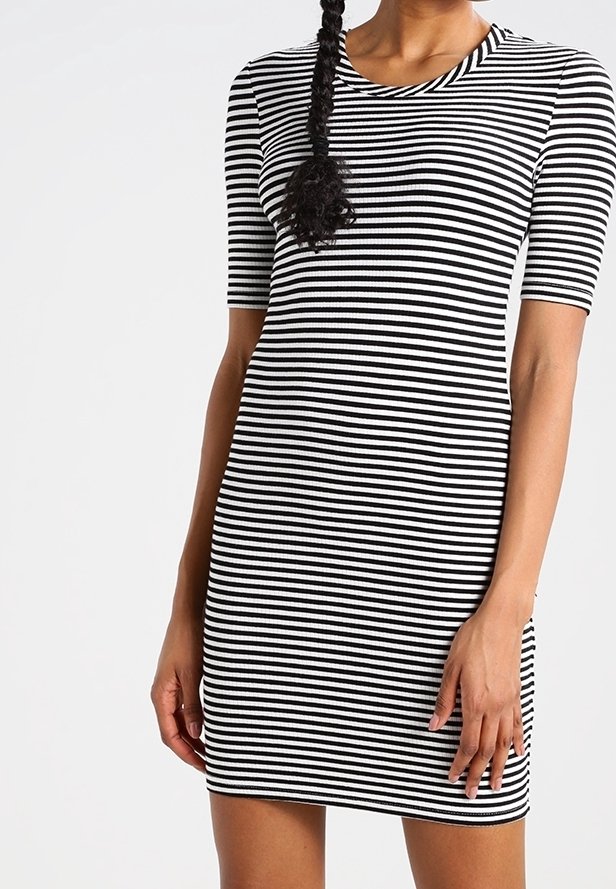 Kvinna iklädd en åtsittande svart och vit randig klänning med ärmar som går ner till armbågarna, stående mot en neutral bakgrund.