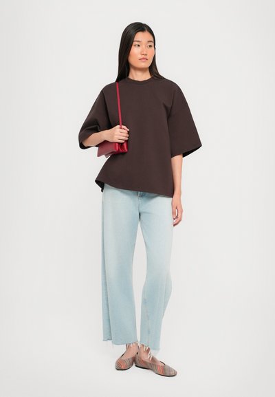 Top corto oversize marrone a maniche corte, jeans a gamba larga blu chiaro con orlo sfrangiato e ballerine a righe colorate. Una borsa rossa è portata in una mano.