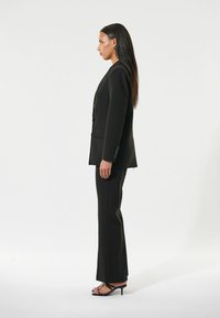 Completo nero su misura con giacca aderente e pantaloni dritti. Tessuto morbido, maniche lunghe, dettagli con bottoni, indossato con tacchi neri a fascia.