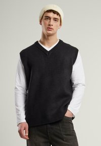 Black sleeveless sweater with a V-neck design, made of a soft material. Worn over a white long-sleeve shirt. Neutral background.