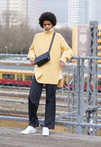Übergroßer gelber Strickpullover mit hohem Kragen, schwarze Umhängetasche, schwarze locker sitzende Hose und weiße Sneaker. Der Hintergrund zeigt eine städtische Landschaft.