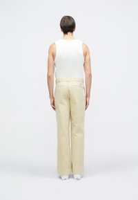 Beżowe spodnie o szerokich nogawkach z widocznymi tylnymi kieszeniami i szczegółem z guzikiem, zestawione z białym, bezrękawnym topem. Gładka faktura tkaniny.