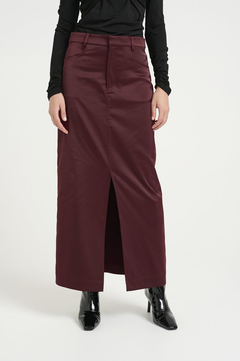 Bordeaux maxi rok met een voorlijn, gemaakt van een gladde stof. Heeft ceintuurlussen en zijzakken, gedragen met zwarte hoge hakken.