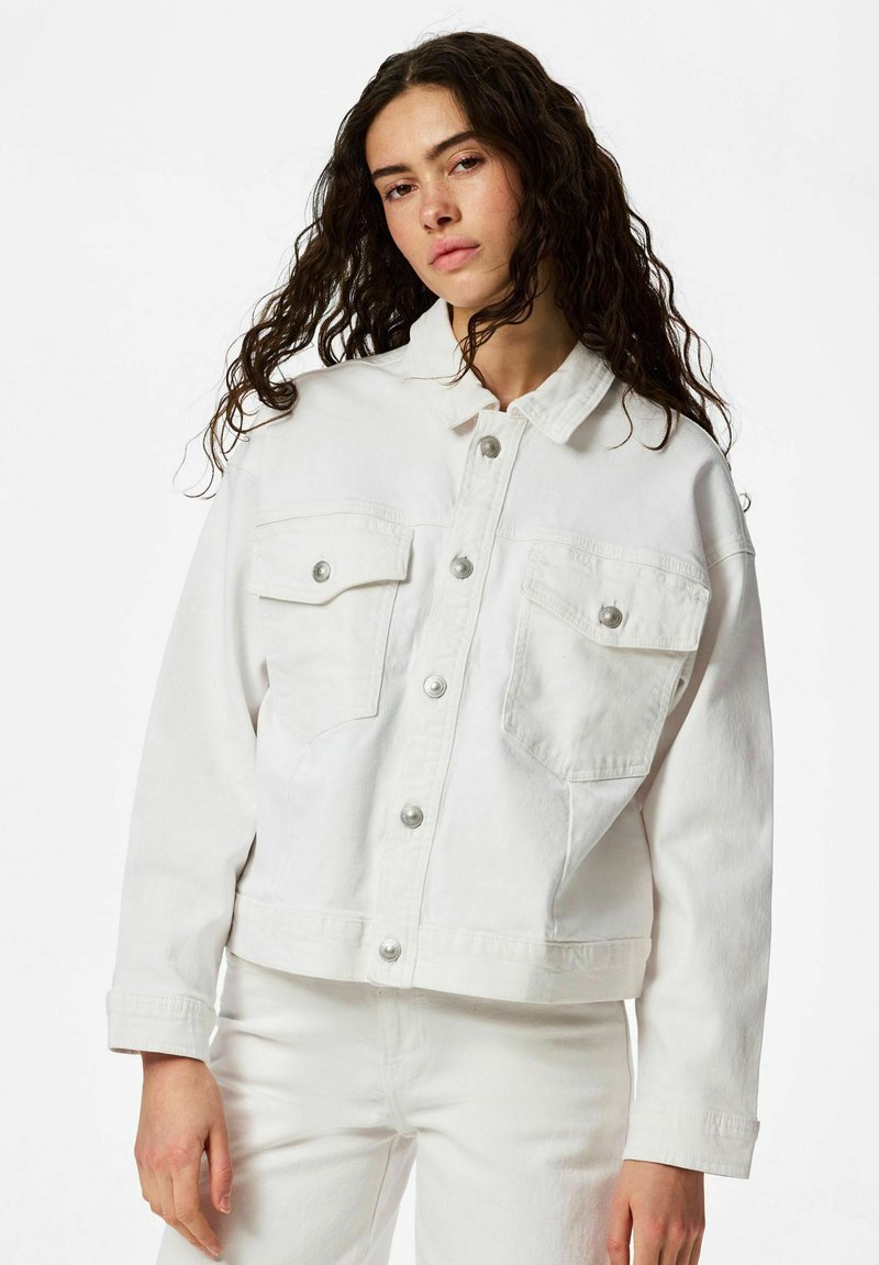 Femme aux longs cheveux bouclés portant une veste en jean blanche surdimensionnée avec des boutons argentés et un pantalon blanc assorti, sur un fond uni.