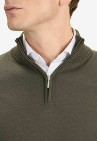 Olivgrön zip-up tröja med strukturerad stickning och kort krage. Har en silverfärgad dragkedja och är lagd över en vit skjorta.