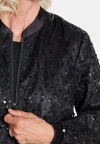 Veste bomber noire avec des sequins brillants, fermeture éclair et poignets côtelés. Texture lisse, tissu léger, éclat subtil des sequins.