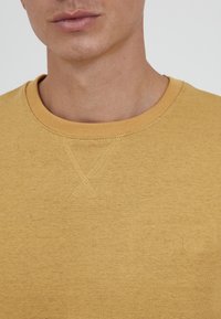 Żółta bluza z okrągłym dekoltem, teksturowaną tkaniną i subtelnym haftem krzyżykowym przy kołnierzu. Posiada małe logo przy krawędzi.