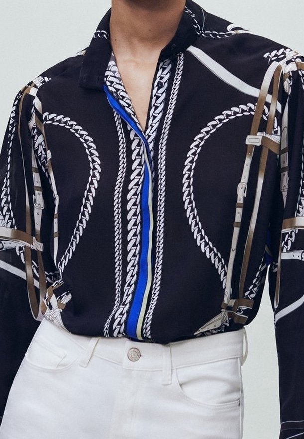 Chemise noire avec motif chaînes blanches et accents bleus, présentant un col à revers et des détails de harnais beige superposés.
