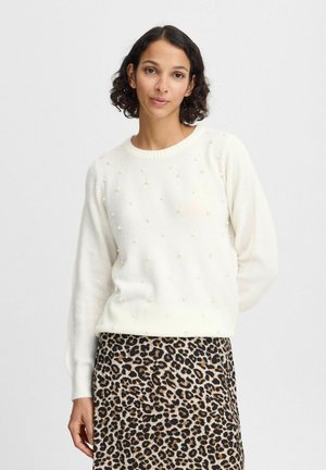 Pull en tricot blanc avec des accents en perles texturées ; col rond et manches longues associés à une jupe imprimée léopard. Fond clair.