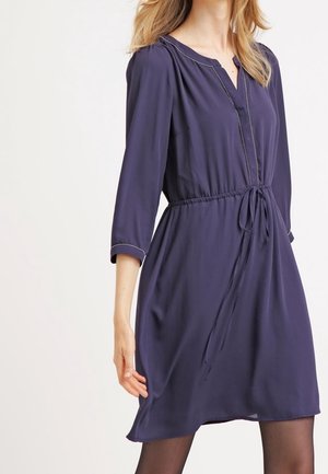 Donna che indossa un vestito blu navy fino al ginocchio con maniche a tre quarti e una cintura in vita, in piedi davanti a uno sfondo chiaro e uniforme.