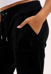 Schwarze Velours-Sweatpants mit einem Kordelzugbund, Seitentaschen und einem gestickten Logo-Detail auf der Tasche. Weicher Stoff und figurbetonte Passform.