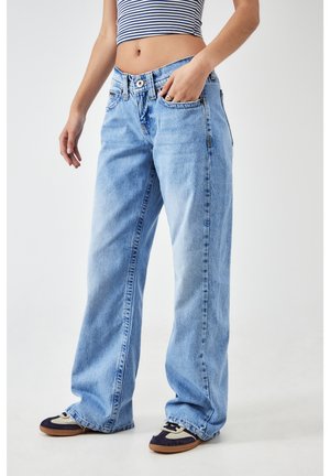 Personne portant un jean bleu clair large, un crop top rayé et des baskets, debout avec une main dans la poche sur un fond uni.