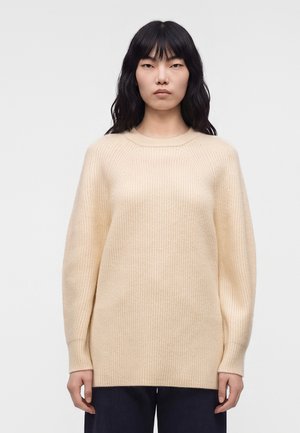 Vrouw met lang zwart haar, die een crèmekleurige geribbelde gebreide trui en donkere broek draagt, staand tegen een effen lichte achtergrond.