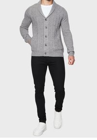 Grauer Cardigan-Pullover mit Zopfmuster, Schalkragen, fünf Knöpfen, langen Ärmeln, kombiniert mit schwarzer Hose und weißen Turnschuhen.