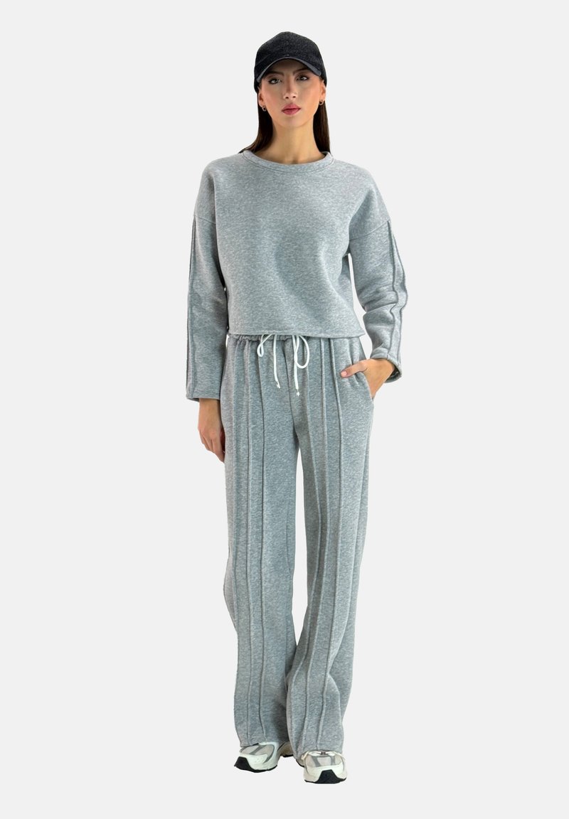 Completo felpa grigia con pantaloni, caratterizzato da un elastico in vita, tasche laterali e righe verticali. Il modello indossa un berretto abbinato.