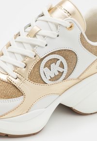 Bílé a zlaté tenisky s texturovaným zlatým svrškem, hladkými zlatými akcenty, bílými šňůrkami a vystouplým logem na boku.