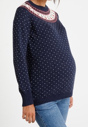Osoba w ciąży ubrana w granatowy dzianinowy sweter z białym i czerwonym wzorem na karczku oraz małymi białymi kropeczkami, w połączeniu z niebieskimi jeansami.