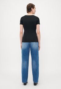 Person mit kurzen lockigen Haaren, die ein eng anliegendes schwarzes T-Shirt, eine blaue gerade Jeans und schwarze Schuhe tr�ägt, steht vor einem schlichten weißen Hintergrund.