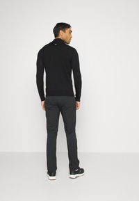 Black long-sleeve sweater with a high collar, paired with dark gray pants. Notable white accents at the shoulder and black footwear.