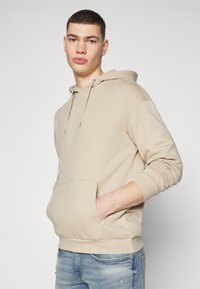 Beige Kapuzenpullover aus weichem Stoff, mit einer Kängurutasche und verstellbarem Kapuzenzug. Getragen mit lässigen hellblauen Jeans, glatte Textur.