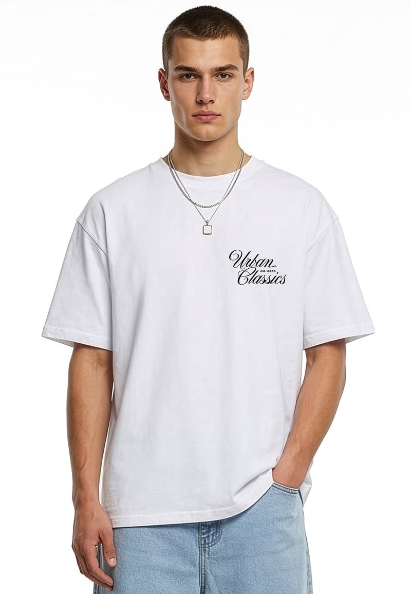 Jeune homme portant un t-shirt blanc "Urban Classics" et un jean bleu clair avec une chaîne argentée, debout avec une main dans la poche.