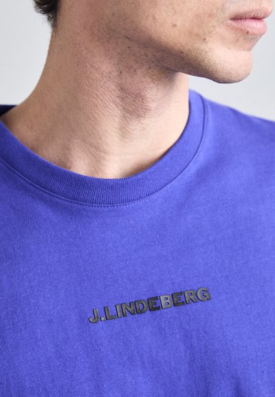 T-shirt violet en coton avec un col rond. Présente un logo noir en relief sur la poitrine, une texture subtile et une coupe standard.