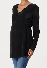 Musta ribattuva äitiystoppi V-päällä, pitkät hihat ja kerrottu vyötärö. Pehmeä pinta ja lentävä muotoilu korostavat siluettia.