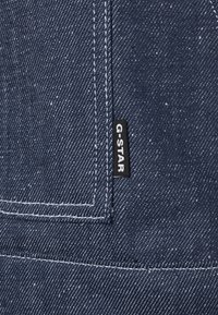 Denimová tkanina v tmavě modré barvě s texturovaným vzhledem, se bílým prošíváním a štítkem G-STAR přišitým na okraji.