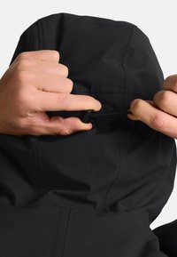 Veste noire avec capuche ; mains ajustant le cordon de serrage. Tissu lisse avec des coutures visibles et une fermeture éclair au niveau du cou.