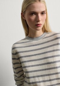 Jeune femme à cheveux blonds longs portant un pull crème à rayures grises horizontales, se tenant contre un mur clair et uni.