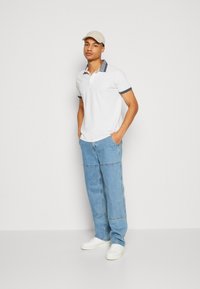 Cămașă polo albă cu guler în dungi bleumarin, pantaloni din denim albastri și adidași albi. Modelul stă cu mâinile în buzunare, purtând o pălărie asortată.