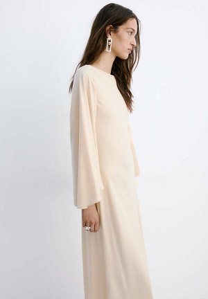 Mujer con cabello largo y oscuro que lleva un vestido suelto de manga larga color crema y pendientes geométricos de plata, de pie frente a un fondo blanco liso.