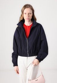 Giacca cropped blu navy con colletto alto, chiusura con zip, vita elasticizzata e maniche voluminose. Indossata sopra una blusa rossa, abbinata a una borsa rosa.