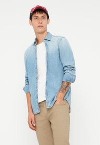 Lys blå denimskjorte, knappet, med oppbrettede ermer. Båret over en hvit t-skjorte og kombinert med khaki bukser. Har et tatovering på underarmen.