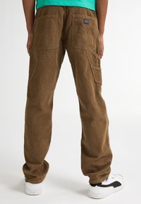 Pantaloni in corduroy marroni con una vestibilità rilassata, dotati di due tasche posteriori e tasche cargo laterali. Indossati con sneakers bianche.