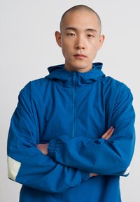 Kék kapucnis dzseki teljes elülső cipzárral és rugalmas mandzsettákkal. Kontrasztos fehér részletekkel a ujjakon, könnyű és sima textúrával.
