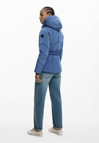 Blauwe pufferjack met een capuchon en elastische tailleband, gekenmerkt door een ruitpatroon en een zwart embleem op de linkerschouder.