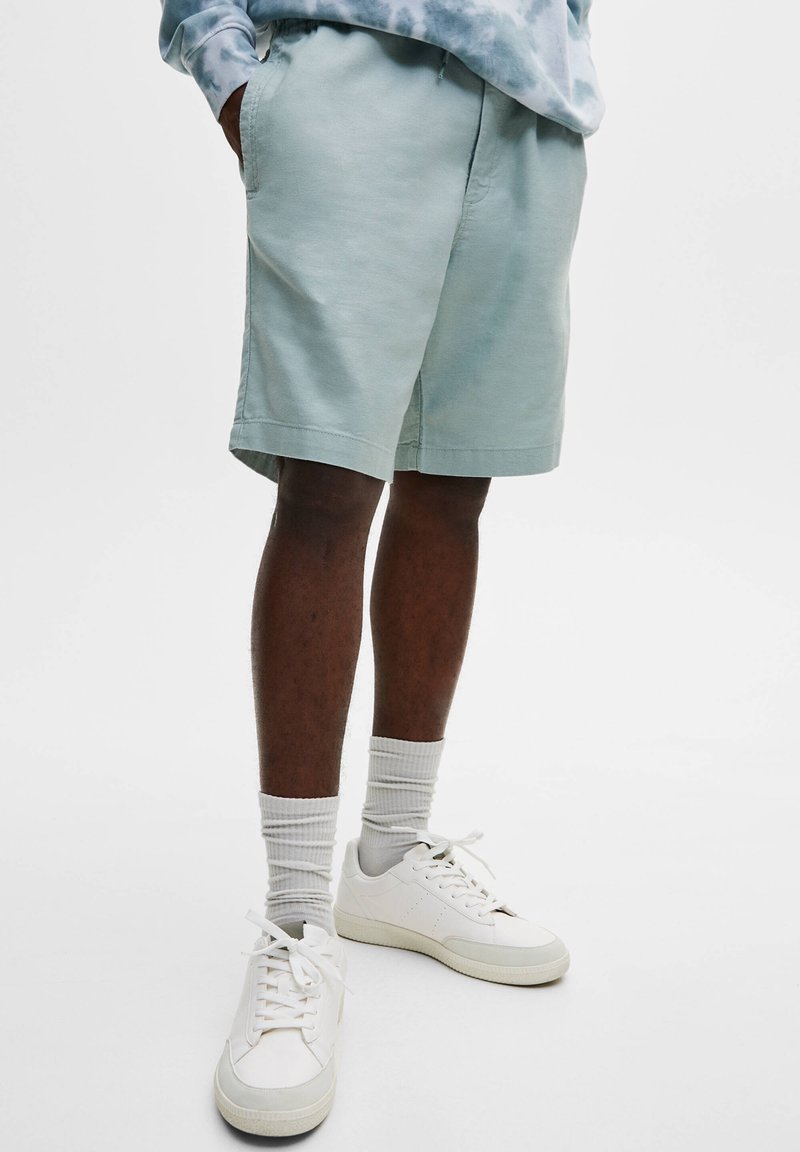 Shorts en coton bleu clair à coupe décontractée, poches latérales, assortis à des baskets blanches et des chaussettes côtelées gris clair. Texture lisse.