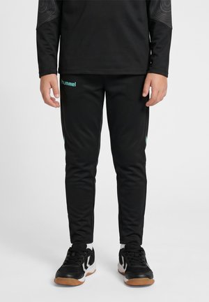 Barn iført sorte sportbukser med blå "hummel" logo og sorte sneakers med hvide såler står mod hvid baggrund.
