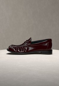 Mocassin en cuir bordeaux foncé brillant avec petit talon et couture style mocassin, présenté sur une surface claire avec un fond neutre.