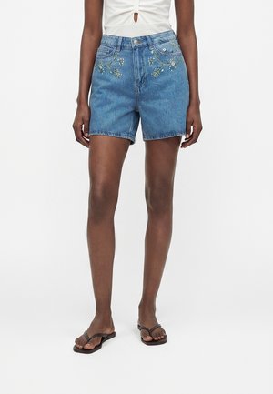 Frau trägt blaue Jeansshorts mit Blumenstickerei, weißes Top und schwarze Flip-Flops, steht vor einem schlichten weißen Hintergrund.