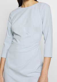 Himmelblå klänning med rund hals och trekvartslånga ärmar. Har en strukturerad tyg med diagonal drapering och en figurnära silhuett.