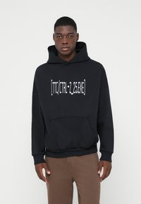 Νεαρός άνδρας ντυμένος με μια μαύρη κουκούλα με μπροστινή τσέπη και λευκό κείμενο "[TTC/CTRL-Z_25.EXE]" σε συνδυασμό με καφέ παντελόνι, ενάντια σε ένα απλό φόντο.