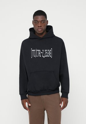 Ung mann iført en svart hettegenser med frontlomme og hvit tekst "[TTC/CTRL-Z_25.EXE]" sammen med brune bukser mot en ensfarget bakgrunn.