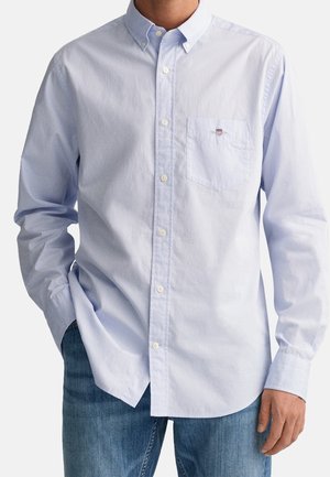 Homme portant une chemise bleu clair à boutons avec poche poitrine et un jean bleu, debout avec une main dans la poche.