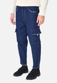 Pantaloni cargo in denim blu scuro con molte tasche, cuciture a contrasto e un fit rilassato. Rifiniti con un orlo sfrangiato e un dettagli con etichetta del logo.