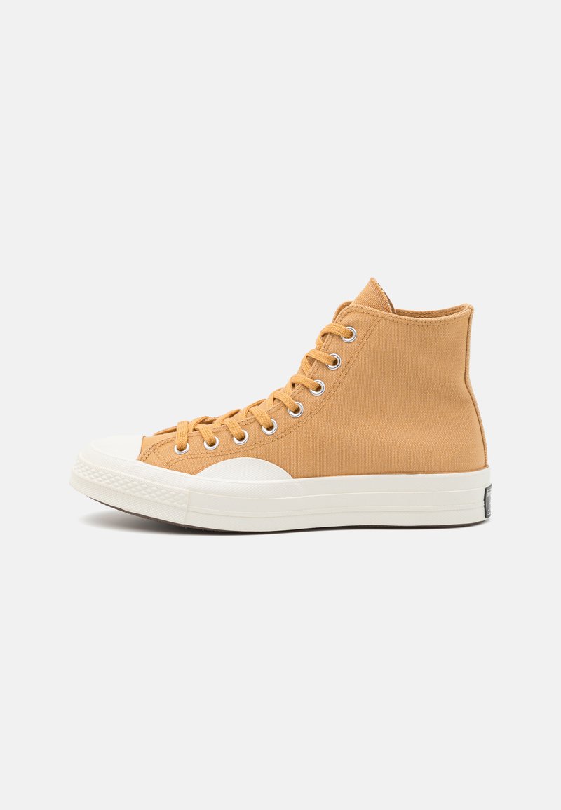 Wysokie trampki w jasnobrązowym płótnie z białym gumowym noskiem i podeszwą. Posiadają okrągłe oczka i płaskie sznurówki. Brak widocznych wzorów.