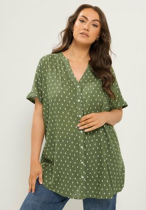 Mujer que lleva una camisa verde de botones de gran tamaño con puntos rectangulares blancos, combinada con vaqueros azules, de pie frente a un fondo liso.