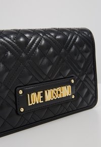 Černá prošívaná kožená peněženka s zlatým logem "LOVE MOSCHINO" na černém koženém štítku, s dekorativními zlatými kovovými detaily.